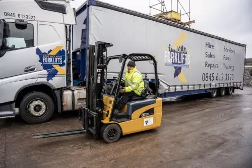 An EMF worker in fluorescent jacket, driving a yellow CAT counterbalance forklift past the East Midlands Forklift lorry